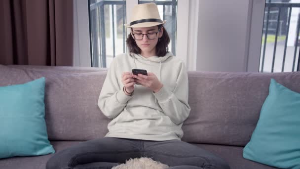 Brunetka dziewczyna w kapeluszu używa telefonu komórkowego w domu na kanapie, tło okna — Wideo stockowe