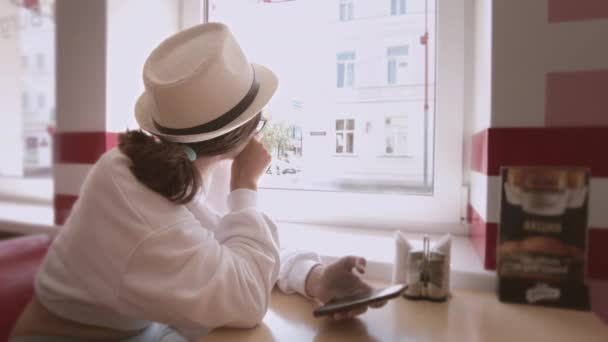 Une fille est assise dans un café, utilisant son téléphone portable, attendant de commander et regardant par la fenêtre — Video