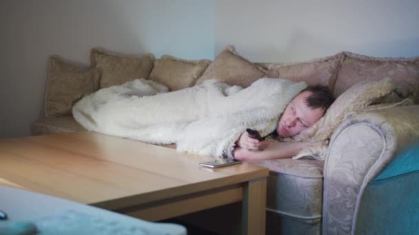 Muž leží v noci na gauči u televize, přepíná kanály dálkovým ovládáním, večerní světlo — Stock video