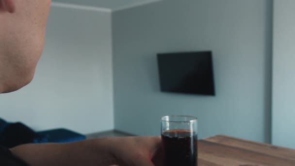 Ένας άνθρωπος βλέπει ένα πρόγραμμα στην τηλεόραση, πίνει ένα ποτό, πλαϊνή άποψη, κοντινό πλάνο — Αρχείο Βίντεο