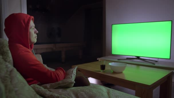 Ein Mann mit Kapuze schaut die Nachrichten im Fernsehen und schaltet die Kanäle mit einer Fernbedienung, sitzt auf der Couch, Chroma-Schlüssel — Stockvideo