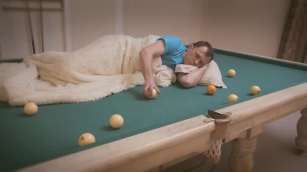 Człowiek jest znudzony, leży na stole bilardowym i toczy piłki na stole, pokryte kocem — Wideo stockowe