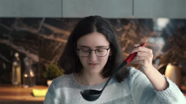 Menina morena degustando a comida deliciosamente cozida da panela com uma concha, olhando para a câmera e sorrindo — Vídeo de Stock