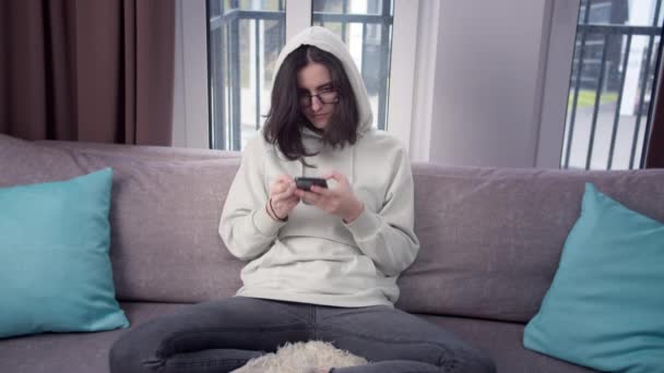 Ragazza in un cappuccio e occhiali utilizzando un telefono cellulare seduto a casa sul divano, sfondo della finestra — Video Stock