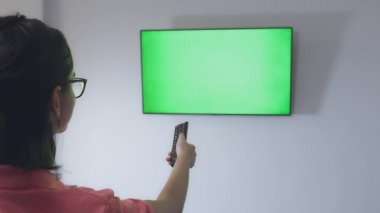 Gözlüklü kız TV 'nin önündeki kanepeye oturur ve uzaktan kumandayla kanalları değiştirir, krom anahtar, yakın plan.