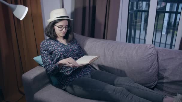 A girl in glasses and a hat is reading while sitting on the couch in the evening with a book in her hands, copy space — Stock Video