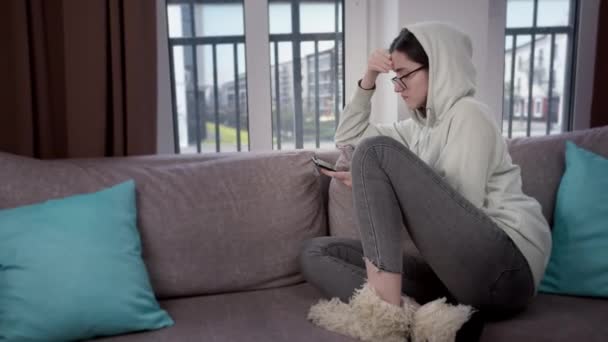 Chica en una capucha y gafas usando un teléfono celular sentado en casa en el sofá, fondo de la ventana, vista lateral — Vídeo de stock