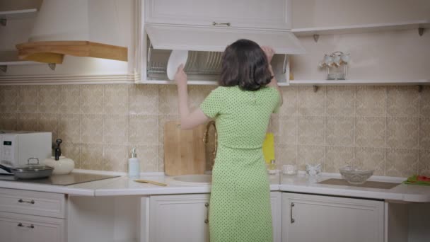 Flicka i en grön klänning sätter saker i ordning i köket skåp, arrangerar rätter, baksida visa — Stockvideo