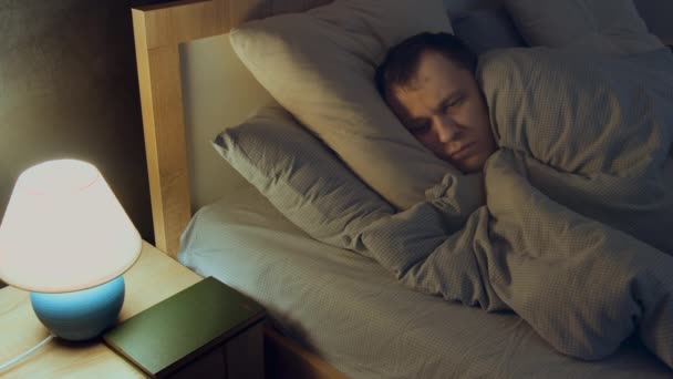 Man lies in bed at night, covered with a blanket, cannot sleep, worried, looks at the lamp — Stock Video