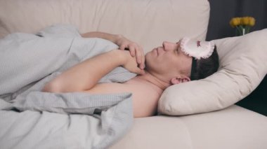 Uyku maskeli genç bir adam battaniyeyle kaplı bir kanepede yatıyor, gözlerine maske takıyor.