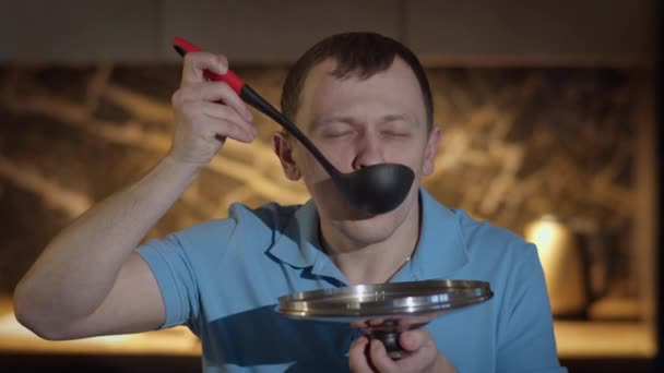 Man met een lepel staat voor de keuken, proeft voedsel uit de lepel, proeft het voedsel — Stockvideo