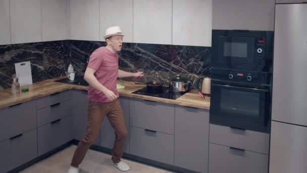 Молодой человек в шляпе танцует на кухне, поет в совок приготовления пищи, хорошее настроение — стоковое видео