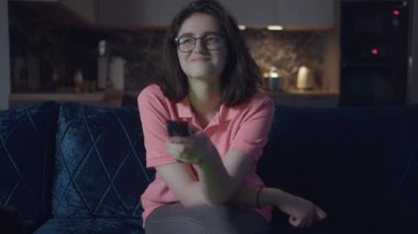Gözlüklü genç kız, gülümsüyor televizyon seyrediyor, kanal değiştiriyor, geceleri kanepede oturuyor.