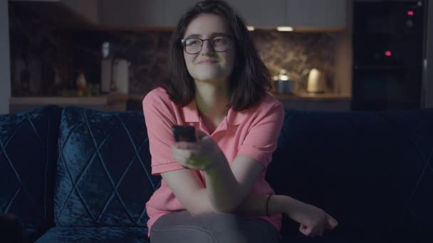 Adolescente en gafas, sonriendo viendo la televisión, cambiando de canal, por la noche sentada en casa en el sofá — Vídeos de Stock