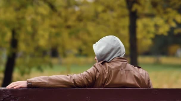 Неизвестный мужчина в капюшоне сидит и ждет на скамейке в осеннем парке — стоковое видео
