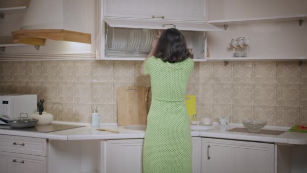 Una chica en un vestido verde pone las cosas en orden en el armario de la cocina, organiza los platos, vista trasera — Vídeo de stock