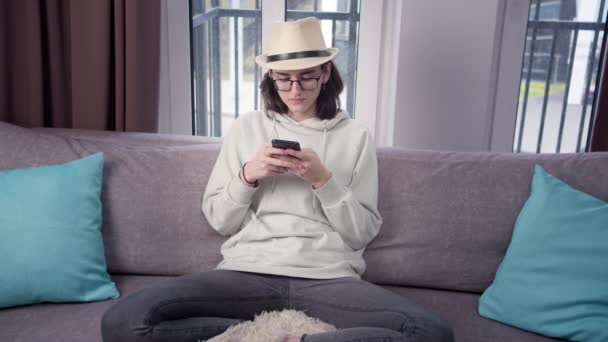 Estudiante chica en un sombrero utiliza su teléfono celular en casa en el sofá, fondo de la ventana — Vídeos de Stock