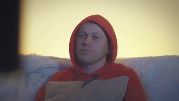 Retrato de un hombre en una capucha mira la televisión por la noche y voltea a través de los canales, primer plano — Vídeos de Stock