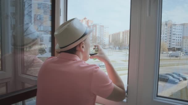 Un hombre con sombrero se para en el balcón mirando a la calle a través de una ventana abierta y bebe — Vídeos de Stock