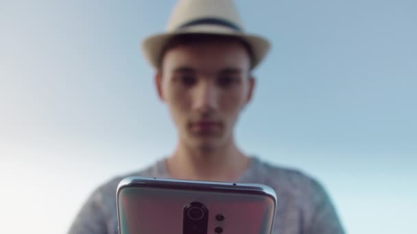 एक नीले आकाश के खिलाफ टोपी में एक आदमी का सिल्हूट अपने हाथों में एक मोबाइल फोन के साथ, रैक फोकस — स्टॉक वीडियो