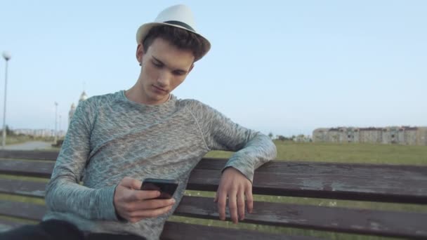 Ritratto di un bel ragazzo con un cappello che siede su una vecchia panchina cittadina con un telefono — Video Stock