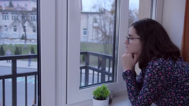 Egy barna, szemüveges lány áll az ablaknál, várja a találkozót, oldalnézetet, másolja a helyet — Stock videók