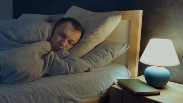 L'uomo ha l'insonnia, non può addormentarsi mentre si trova a letto la notte, la lampada è sul comodino — Video Stock