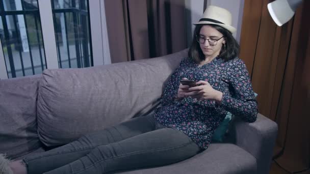 En tenåring med briller og hatt leser beskjeder på mobilen mens hun sitter på sofaen om kvelden. – stockvideo