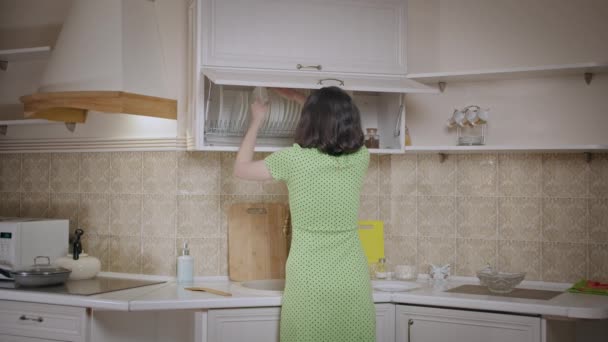 A ruhás lány rendet tesz a konyhaszekrényben, elrendezi az edényeket, visszapillantás — Stock videók
