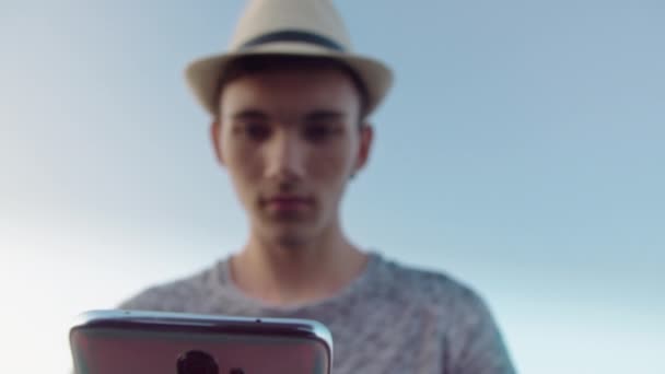 Silhouette floue d'un gars coiffé d'un chapeau contre un ciel bleu avec un téléphone portable dans les mains — Video