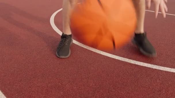 Pernas homem praticando habilidades de bola, rolando uma bola de basquete entre as pernas em uma quadra ao ar livre, rastreamento de câmera — Vídeo de Stock