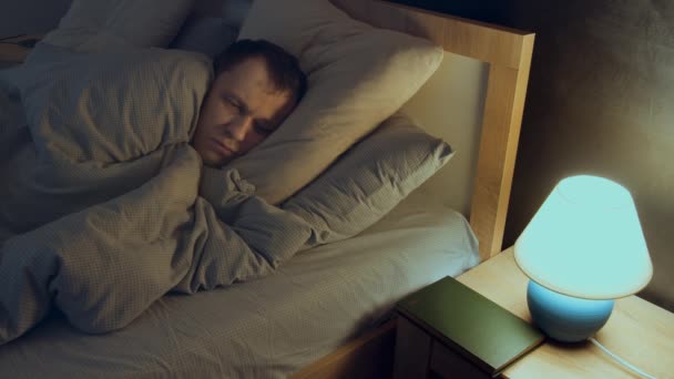 Uykusuzluk çeken bir adam, geceleri yatakta yatarken uyuyamaz, lamba komodinin üzerinde. — Stok video