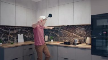 Şapkalı bir adam ocakta yemek hazırlıyor, evinde elinde kepçeyle dans ediyor ve şarkı söylüyor.