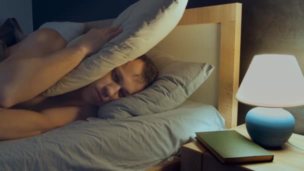 Man kan inte somna liggande i sängen på natten på grund av buller, täcker sitt huvud med en kudde — Stockvideo