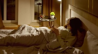 Gözlüklü bir kız gece yatağa uzanıyor, battaniyeyle örtülü, mobil uygulama kullanıyor, gece lambası kullanıyor, kamera takibi yapıyor.