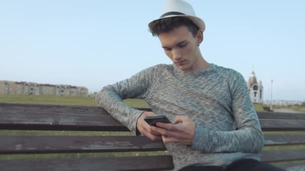 Un tizio con un cappello e un orecchino nell'orecchio, che siede su una vecchia panchina della citta 'con un telefono, legge messaggi, segue le telecamere — Video Stock