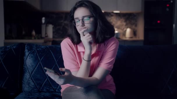 Menina com óculos senta-se na frente da TV à noite e lê mensagens em seu telefone, vista frontal — Vídeo de Stock