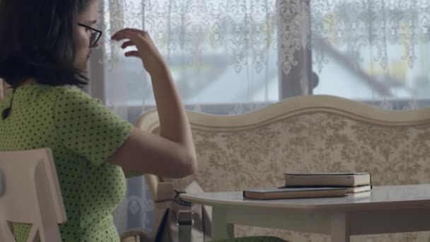 A girl with glasses and a green dress sits at her desk and reads messages on her phone, side view — Vídeo de Stock