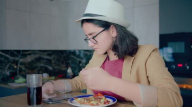Şapkalı ve gözlüklü esmer kız, yemek yiyor ve mobil uygulama