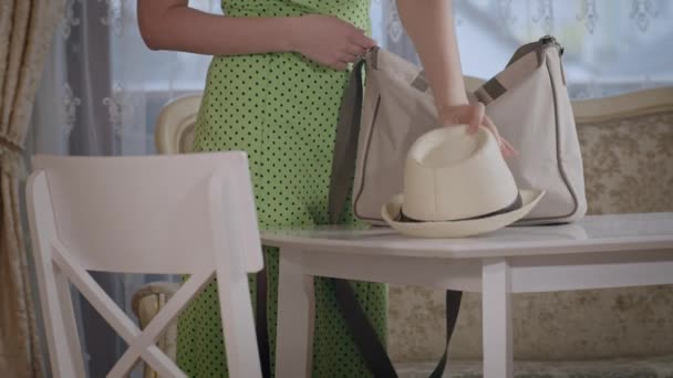 Дівчина в зеленій сукні кладе сумку на стіл і кладе капелюх, виймає підручники, блокнот і ручку з сумки — стокове відео