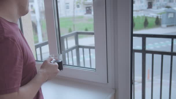 Чоловік з чашкою кави дивиться у вікно квартири на вулиці — стокове відео
