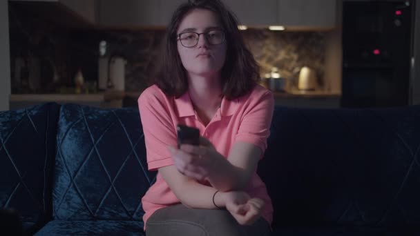 Sad girl with glasses at home in the evening watching TV while sitting on the couch, kitchen background, front view — Αρχείο Βίντεο