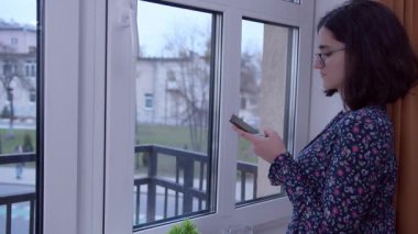 Girl with a mobile phone in her hands standing at the window, side view, copy space
