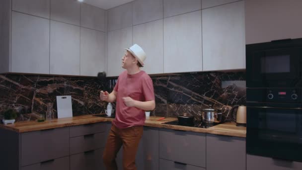 Fiatal férfi kalapban táncol és énekel egy merőkanállal a kezében a konyhában főzés közben — Stock videók