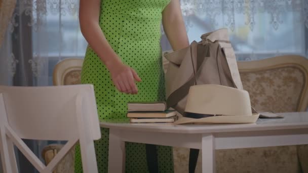 Una chica en un vestido pone una bolsa en la mesa y se pone el sombrero, saca libros de texto, un cuaderno y un bolígrafo de la bolsa — Vídeo de stock