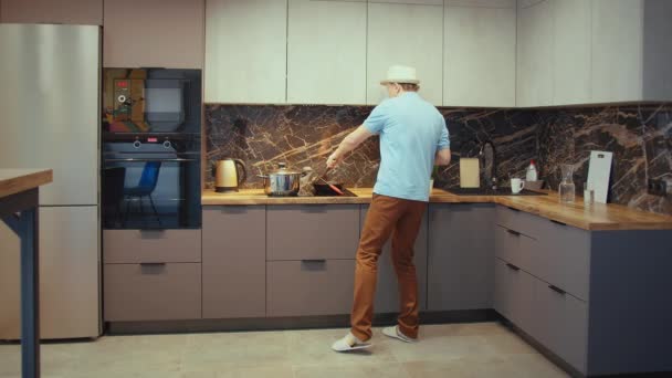 Fiatal férfi kalapban szórakozás a konyhában főzés közben, tánc és éneklés egy merőkanállal a kezében — Stock videók
