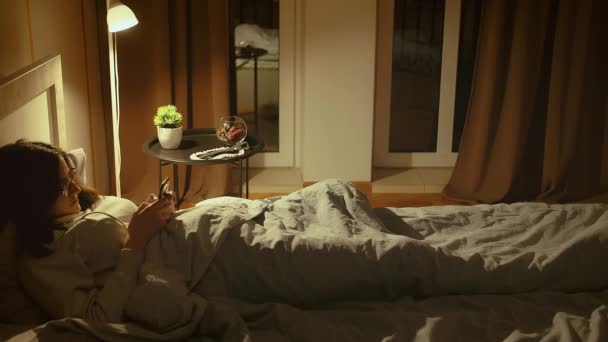 Brunetka w okularach leży w łóżku w nocy, przykryta kocem, korzysta z aplikacji mobilnej, lampy wieczornej, śledzenia kamer — Wideo stockowe