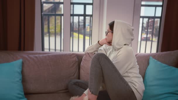 Mädchen mit Kapuze schaut nachdenklich aus dem Fenster, während sie auf dem Sofa sitzt, kopiert Raum — Stockvideo