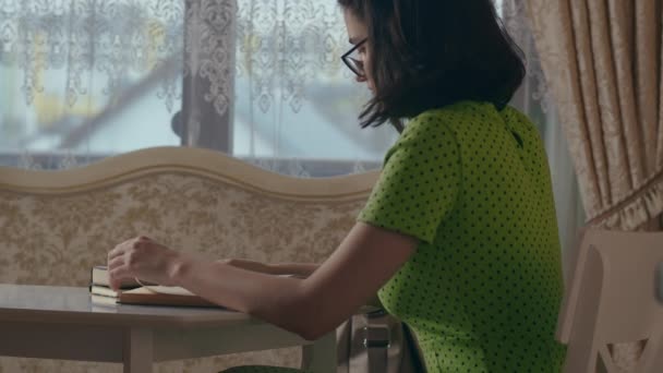 Gözlüklü ve yeşil elbiseli bir kız masada oturmuş kitap okuyor. — Stok video