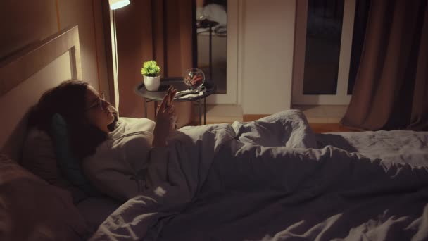 Menina de óculos fica na cama à noite, coberto com um cobertor, usa um aplicativo móvel, luz da noite de uma lâmpada — Vídeo de Stock